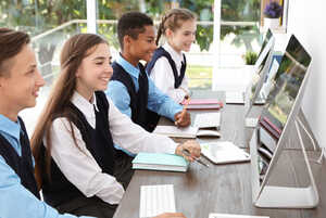 Four students at computers, engaged and smiling, with notebooks and stationery on the table.