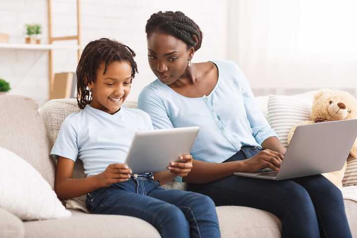 Mother and child sat next to each other, child is grinning and holding a tablet, while the mother is looking over their shoulder to see what they are doing.